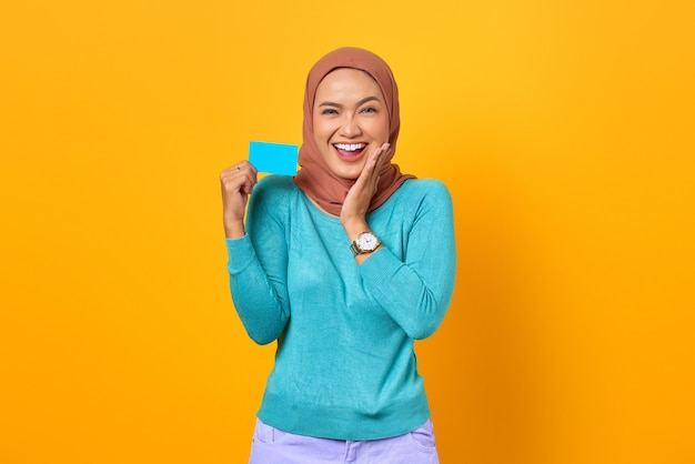 Happy young Asian woman holding credit card and touch cheeks on yellow background