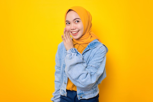 Happy young Asian woman in jeans jacket hand on mouth telling secret and whispering gossip isolated over yellow background