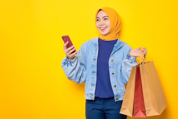 청바지 재킷을 입은 행복한 젊은 아시아 여성이 휴대전화와 쇼핑백을 들고 노란색 배경에 격리된 복사 공간을 옆으로 바라보고 있습니다.