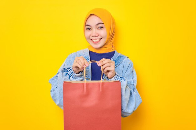 Happy young Asian woman in jeans jacket holding shopper bags and smiling to camera isolated over yellow background