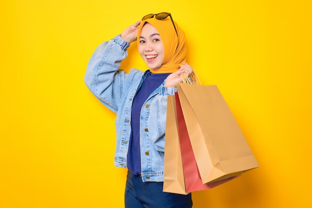 Happy young Asian woman in jeans jacket holding shopping bags and touching sunglasses looking at camera isolated over yellow background