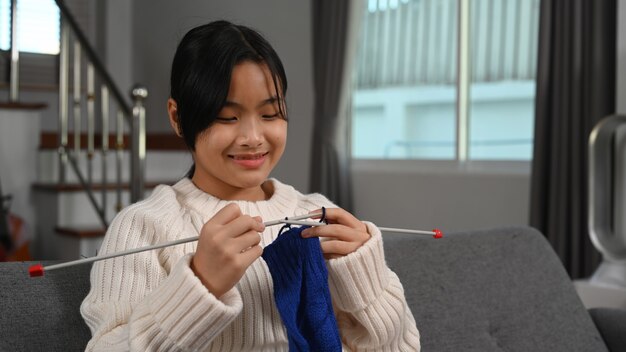 Happy young asian woman knitting blue sweater while sitting in living room.