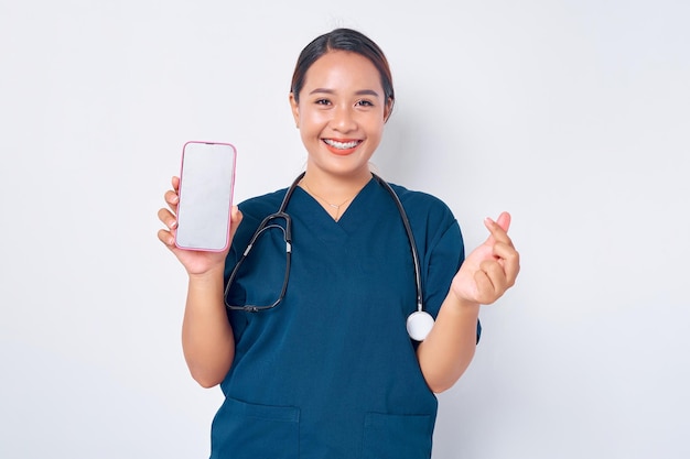 Happy young Asian woman professional nurse working wearing a blue uniform showing Korean heart love gesture and mobile phone with blank screen isolated on white background Healthcare medicine concept