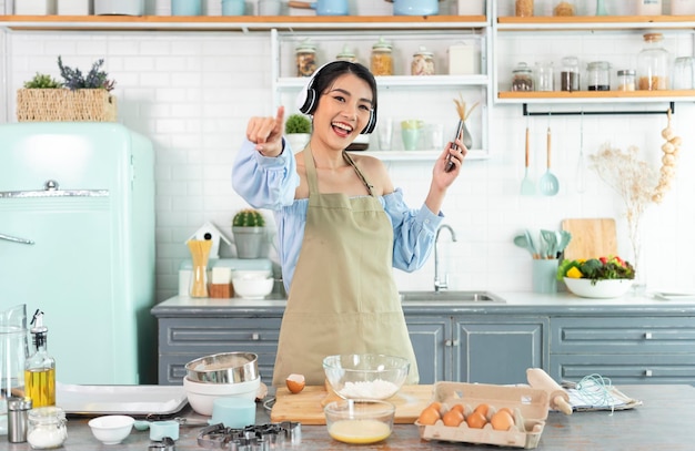 Happy young asian woman raise smartphone fist good mood wear headphones listen to music in kitchen