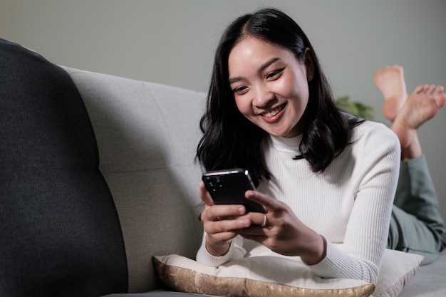 행복한 젊은 아시아 여성은 집에서 편안한 소파에서 휴식을 취하고 스마트폰에서 문자 메시지를 보내며 미소 짓는 소녀는 휴대전화를 사용하여 온라인 메시지를 채팅합니다.
