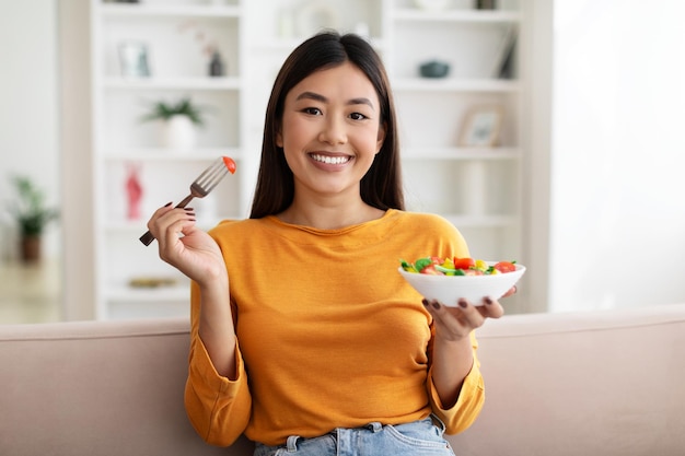 サラダを食べてソファに座って幸せな若いアジアの女性