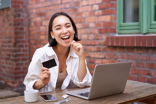 Happy young asian woman sitting near laptop holding credit card\
paying bills shopping online contact