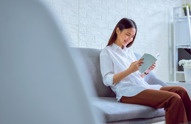 Giovane donna asiatica felice che si siede sul sofà e sul libro di lettura nel suo salone, tempo di vacanza.