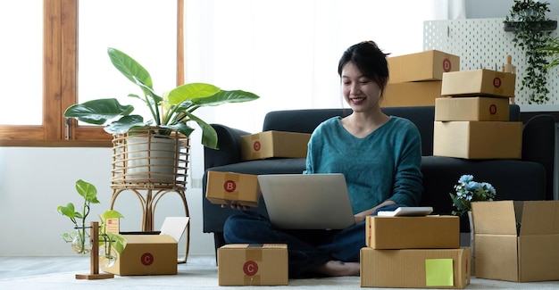 Happy young asian woman startup small business freelance holding parcel box and computer laptop and sitting on floor Online marketing packing box delivery concept