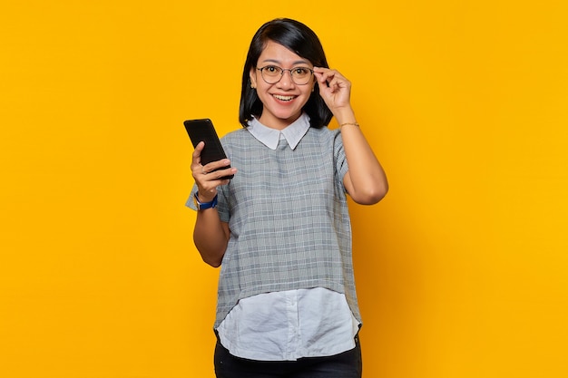 Happy young Asian woman touching glasses and holding mobile phone on yellow background