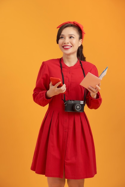 Happy young asian woman tourist holding phone passpoer camera and standing over orange background