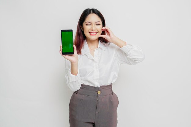 A happy young Asian woman wearing a white shirt showing shapes heart gesture expresses tender feelings while showing copy space on her phone