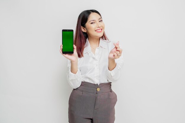 A happy young Asian woman wearing a white shirt showing shapes heart gesture expresses tender feelings while showing copy space on her phone