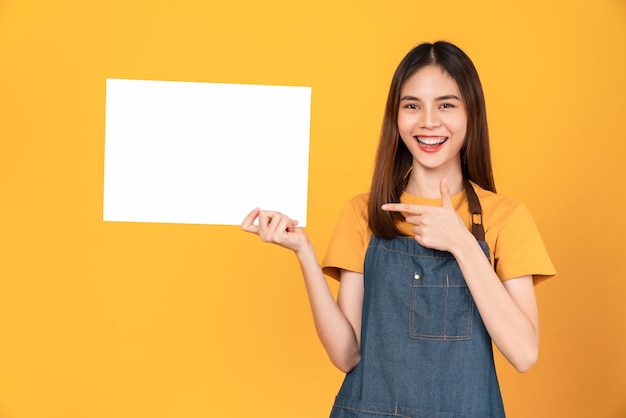 Felice giovane donna asiatica indossa un grembiule e tiene un foglio bianco con la faccia sorridente e guardando lo sfondo arancione. per insegne pubblicitarie.