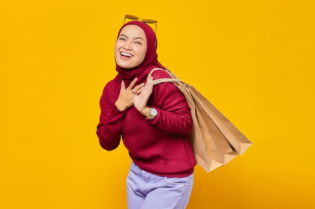 Счастливая молодая азиатская женщина с рукой на сундуке и держащая хозяйственные сумки на желтом фоне