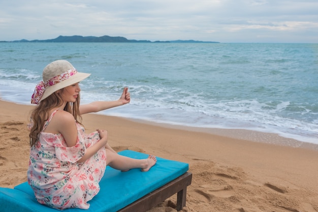 Happy young Asian woman with hat relaxing on beach chair and doing finger heart.