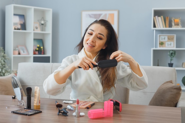 happy young asian woman with towel in bathrobe sitting at the dressing table at home interior touching her hair brushing her hair doing morning makeup routine