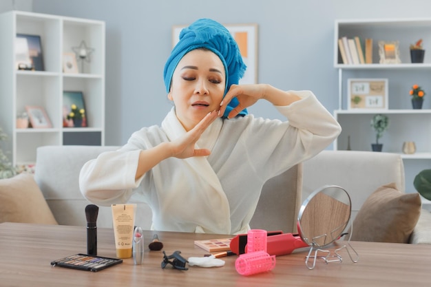 Счастливая молодая азиатка с полотенцем на голове сидит за туалетным столиком в домашнем интерьере, смотрит в зеркало, делает массаж лица, делает утренний макияж