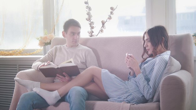 Happy young attractive couple sitting together on the couch the guy reads a book the girl use phone