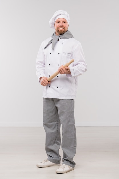 Happy young bearded chef in uniform holding wooden rolling-pin while standing in front of camera in isolation