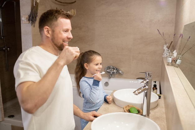 Happy young bearded father and his cute little cheerful daughter holding toothbrushes by their teeth while standing in bathroom