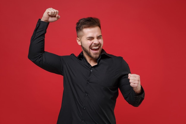 Photo happy young bearded guy 20s in classic black shirt posing isolated on bright red background studio portrait. people sincere emotions lifestyle concept. mock up copy space. clenching fists like winner.