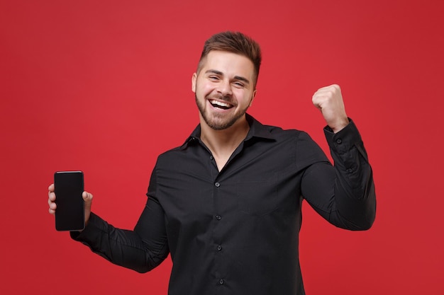 Happy young bearded guy 20s in classic black shirt posing isolated on red wall background. People lifestyle concept. Mock up copy space. Hold mobile phone with blank empty screen doing winner gesture.
