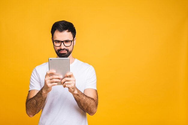 Happy young bearded man in casual standing and using tablet isolated over yellow background.