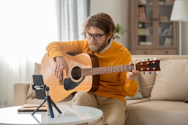 Happy young bearded man in eyeglasses, jeans and jumper looking in smartphone camera during online lesson of guitar playing