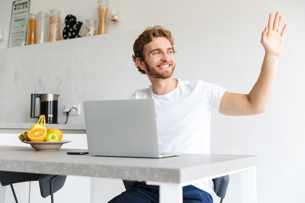  happy young bearded man at the table at home talking using laptop computer waving.