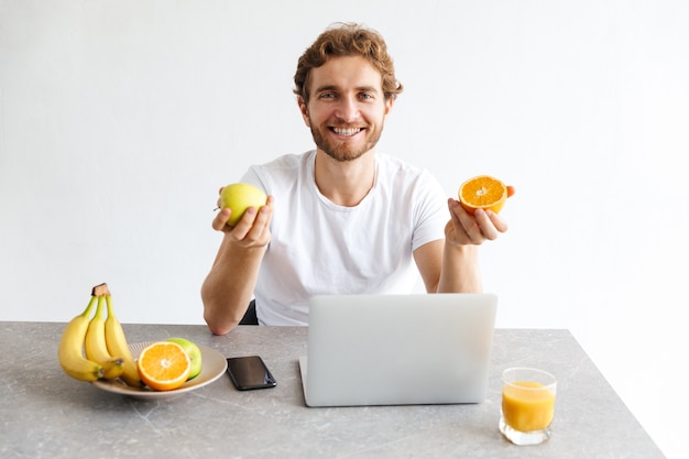  happy young bearded man at the table at home using laptop computer holding fruits.