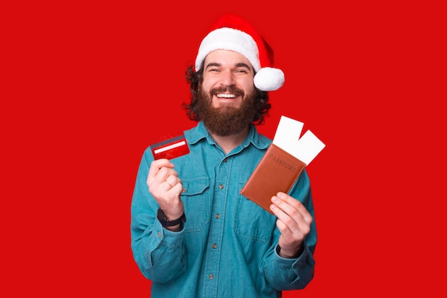 산타 클로스 모자를 쓰고 빨간 신용 카드와 비행기 티켓이 있는 여권을 보여주는 수염 난 행복한 젊은이