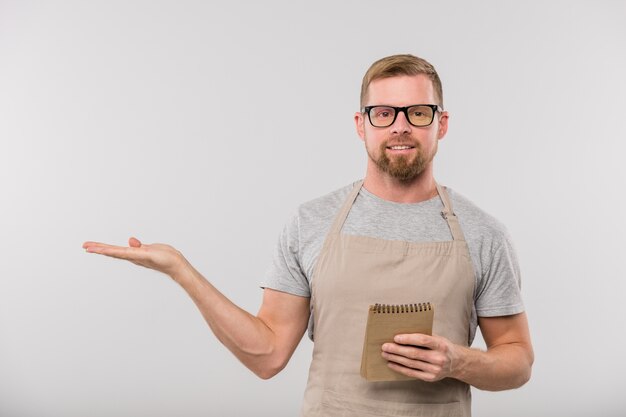 Happy young bearded waiter in apron and eyeglasses showing aside while looking at you in isolation