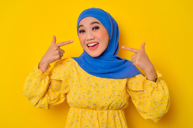 Happy young beautiful Asian Muslim woman showing perfect white teeth isolated over yellow background