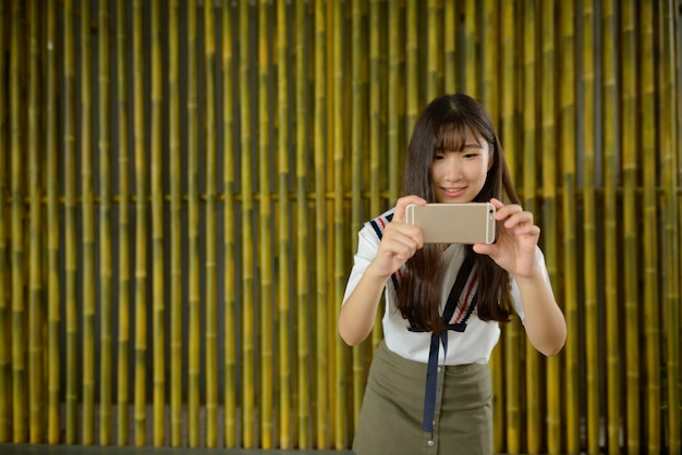 Счастливая молодая красивая азиатская девочка-подросток фотографирует с телефоном у бамбукового забора