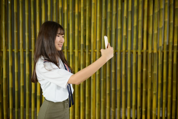 Happy young beautiful asian teenage girl taking selfie against bamboo fence