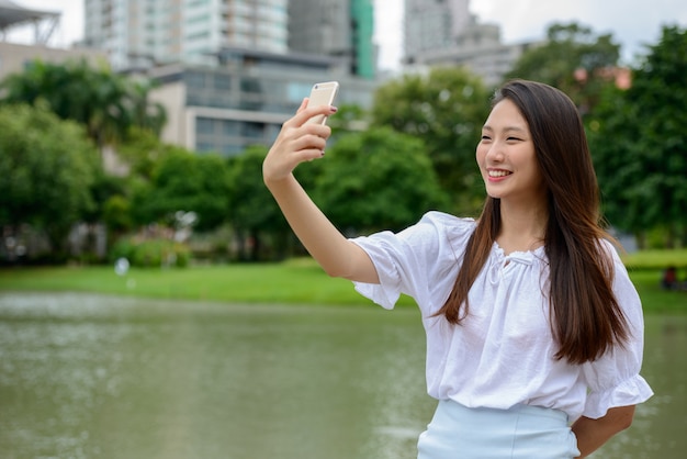 Счастливая молодая красивая азиатская подростковая женщина принимая selfie на парк