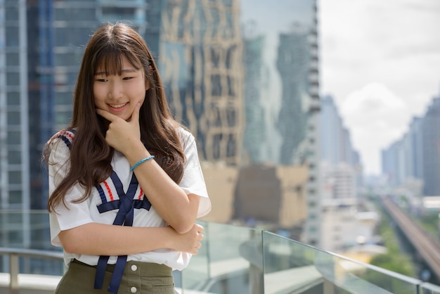 Счастливая молодая красивая азиатская подростковая женщина думая против взгляда города