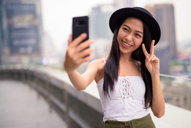 Felice giovane bella donna turistica asiatica prendendo selfie contro la vista della città