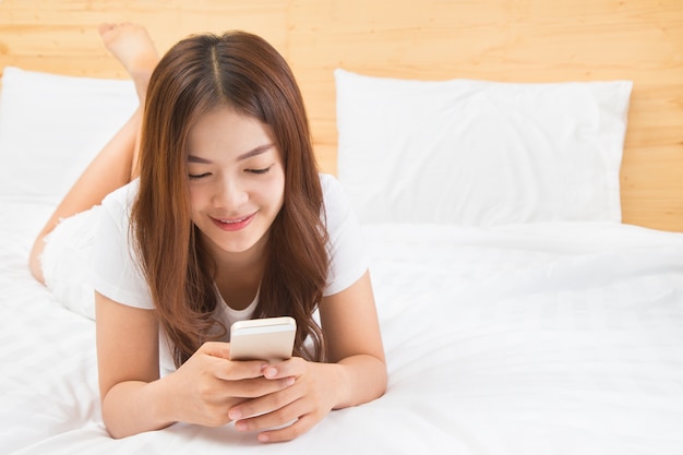 그녀의 스마트 폰을 사용하여 그녀의 흰색과 밝은 조명 침실에 누워 행복 한 젊은 아름 다운 아시아 여자