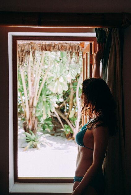 Happy young beautiful female stands at opened window in her bungalow and looks outside to tropical garden. travel and vacation concept