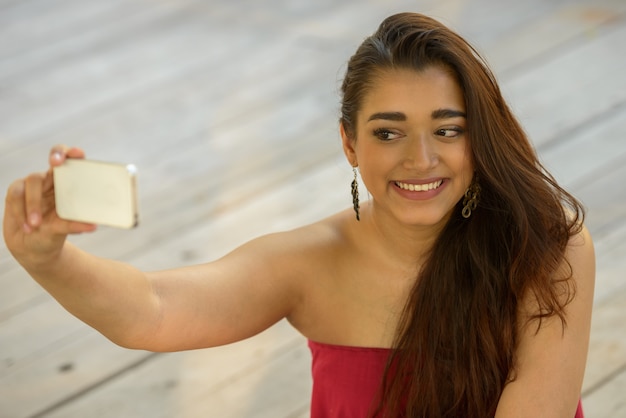 Happy young beautiful Indian woman taking selfie while sitting on wooden floor