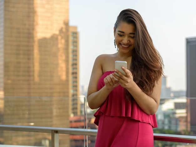 Happy young beautiful Indian woman using phone against view of the city