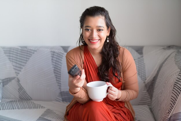 Happy young beautiful Indian woman with coffee laughing while watching tv at home