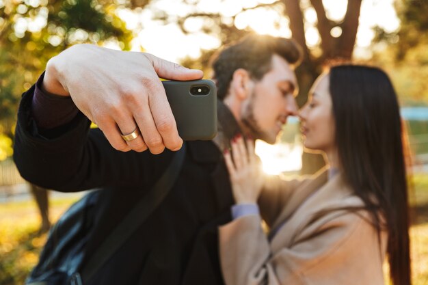 Фото Счастливая молодая красивая любящая пара, позирующая гуляя на открытом воздухе в парке природы, принимает селфи поцелуями мобильного телефона.