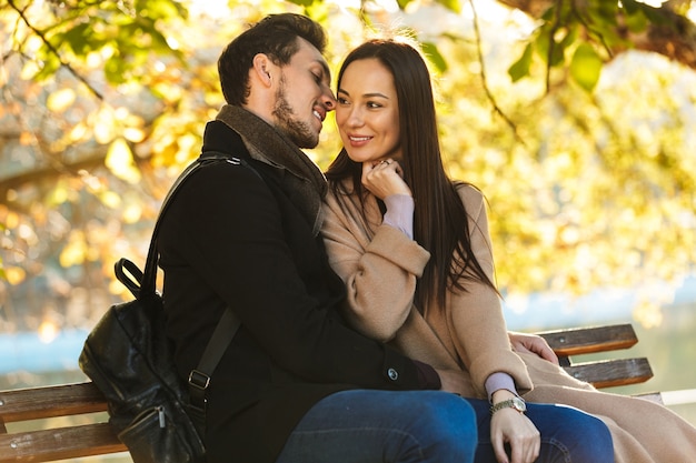 幸せな若い美しい愛情のあるカップルは、ベンチのキスに座って公園の自然の中で屋外を歩いてポーズをとる。