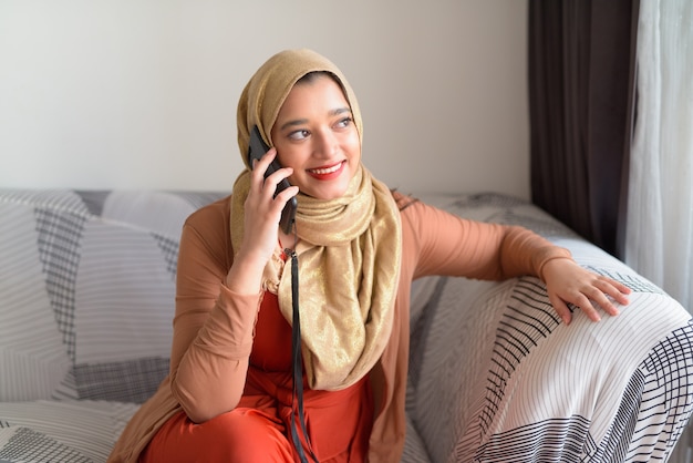 Happy young beautiful Muslim woman talking on the phone in the living room at home