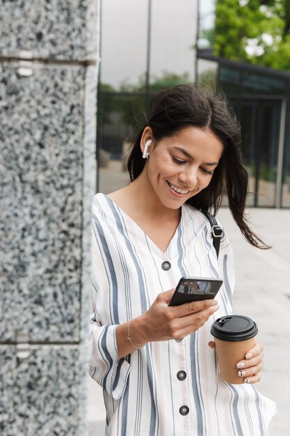 Felice giovane bella donna d'affari in posa all'aperto fuori a piedi chiacchierando con il cellulare bevendo caffè ascoltando musica con gli auricolari.