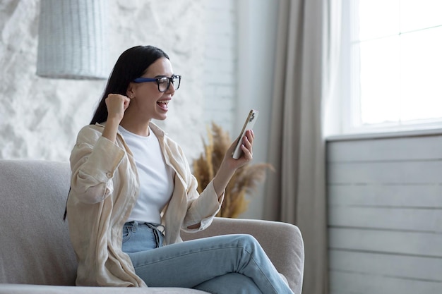 Happy young beautiful woman in glasses sitting on sofa at home with phone in hands celebrates a