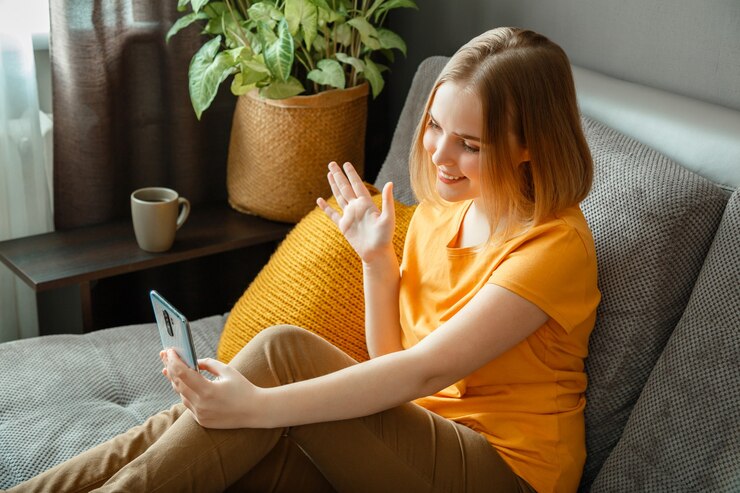 फोटो happy young beautiful woman greets, wave by hand greeting, talking by video call using smartphone while sitting in living room. teen girl make video call communication in home interior.